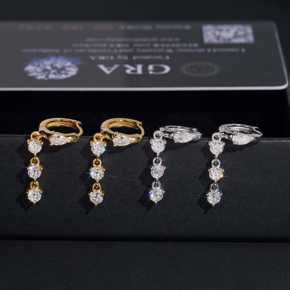 Iced out pear cut and 3-stone drop dangle huggie hoop moissanite earrings in gold and silver in front of GRA card   
