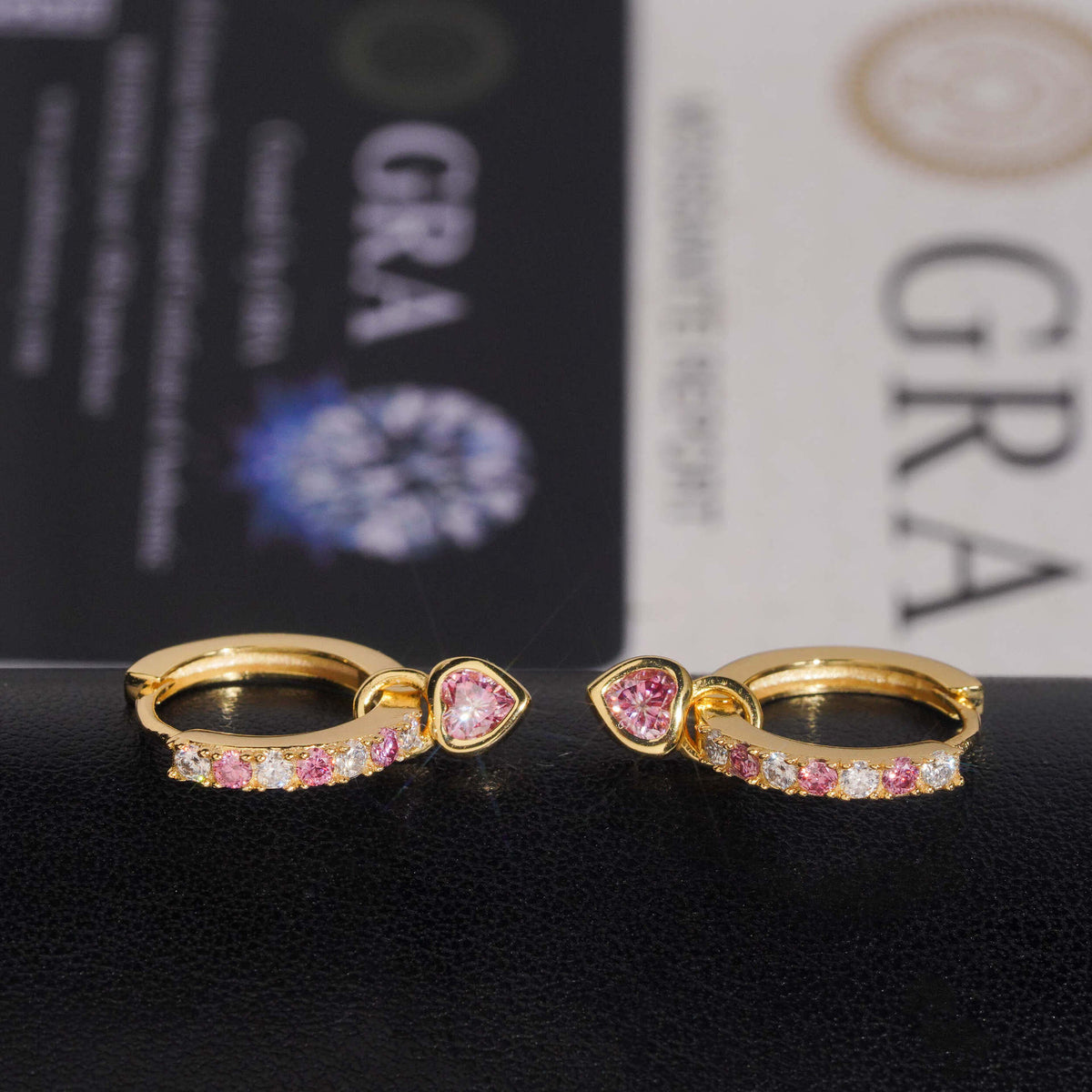 Pair of yellow gold iced out pink heart dangle huggie hoop moissanite earrings resting on GRA report card
