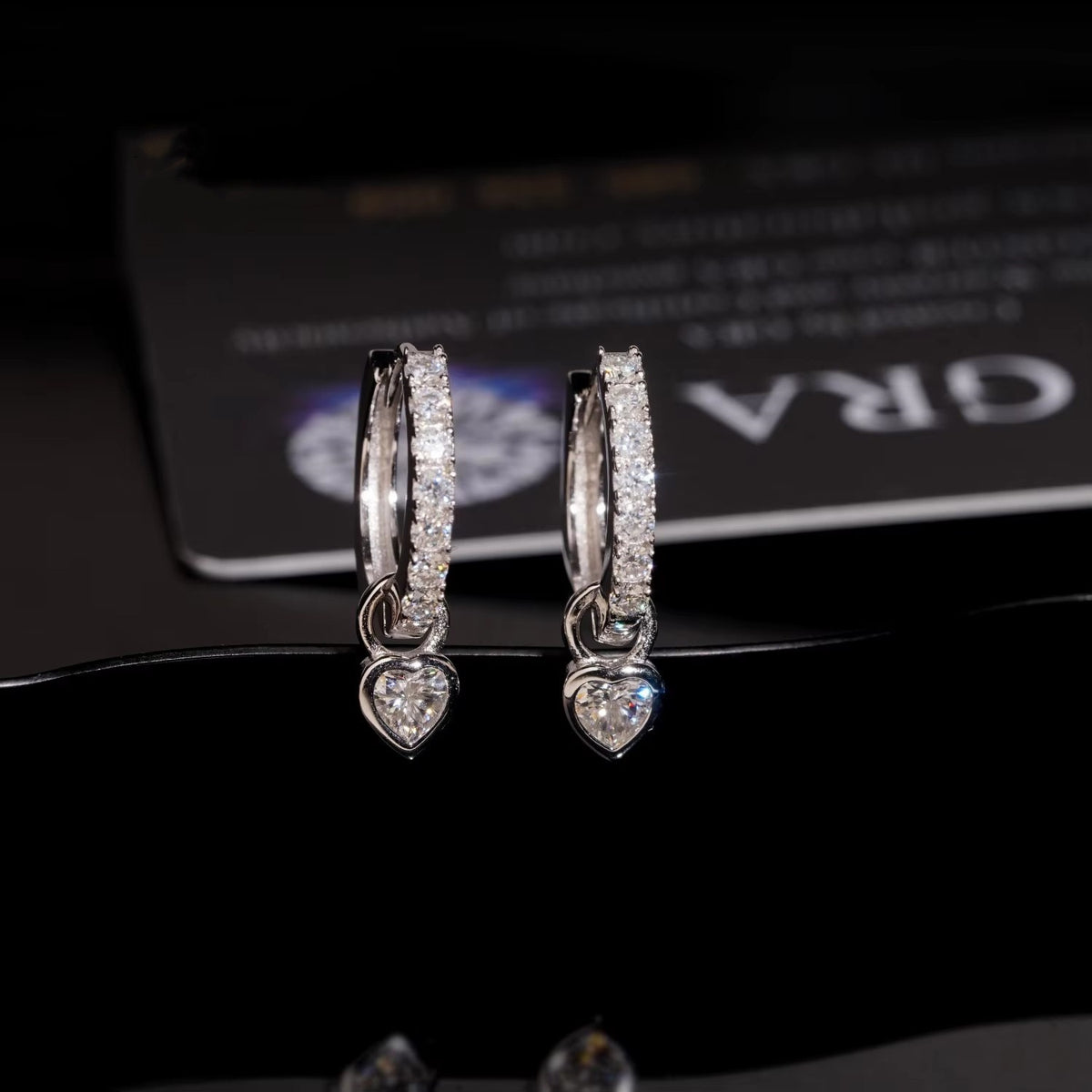 Silver iced out pink heart dangle huggie hoop moissanite earrings in front of GRA certificate card