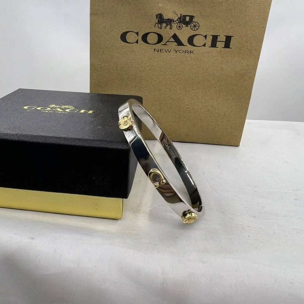 A Coach bangle bracelet with a polished silver-toned band and gold-tone logo charms, displayed in front of a black Coach box and a brown Coach box on a light surface.