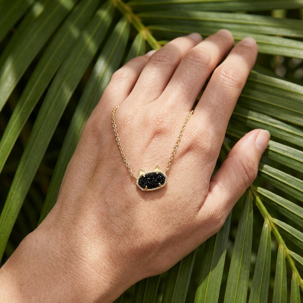 The Kendra Scott Elisa cat gold and black necklace held on a hand against a tropical palm leaf background for scale.