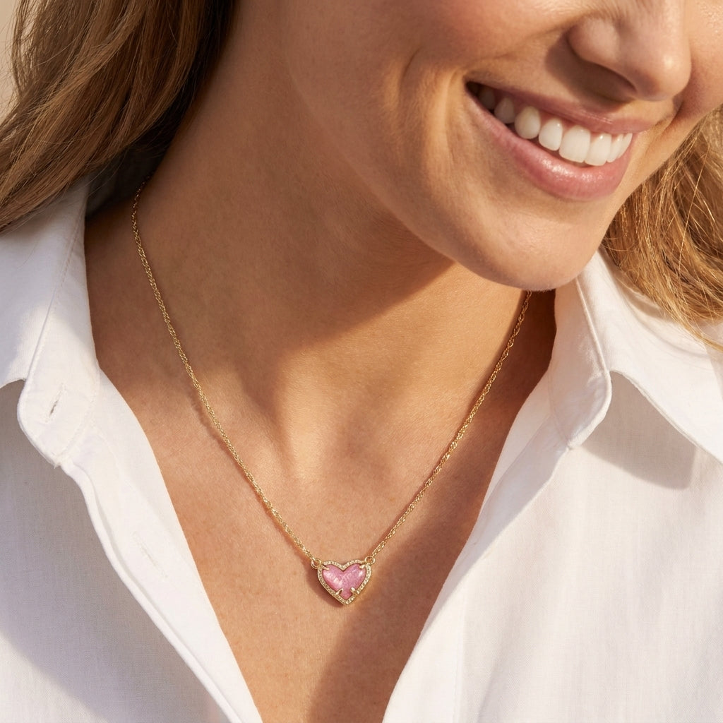 Close-up of a model wearing the gold Kendra Scott Ari pink heart pendant necklace with a white blouse.