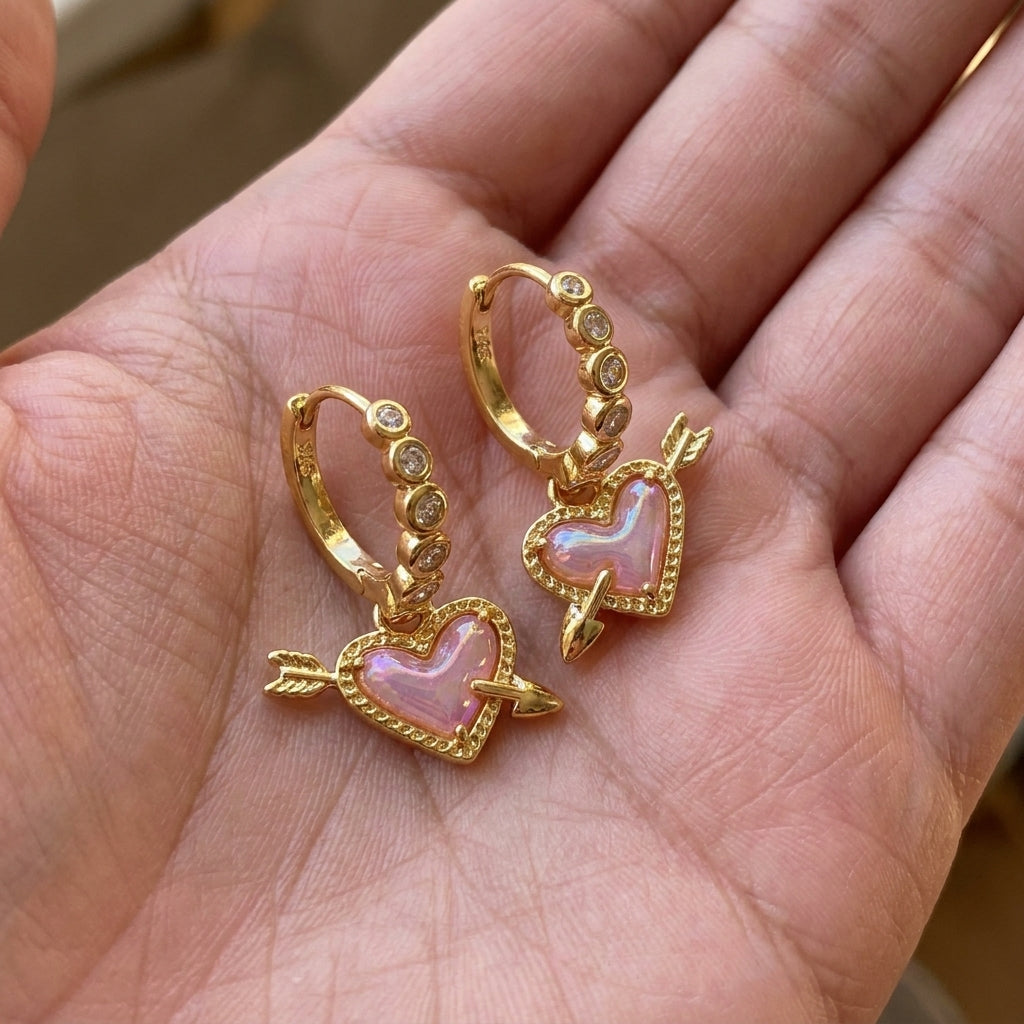 Hand holding the gold Kendra Scott Ari heart arrow huggie earrings to show natural light reflection.