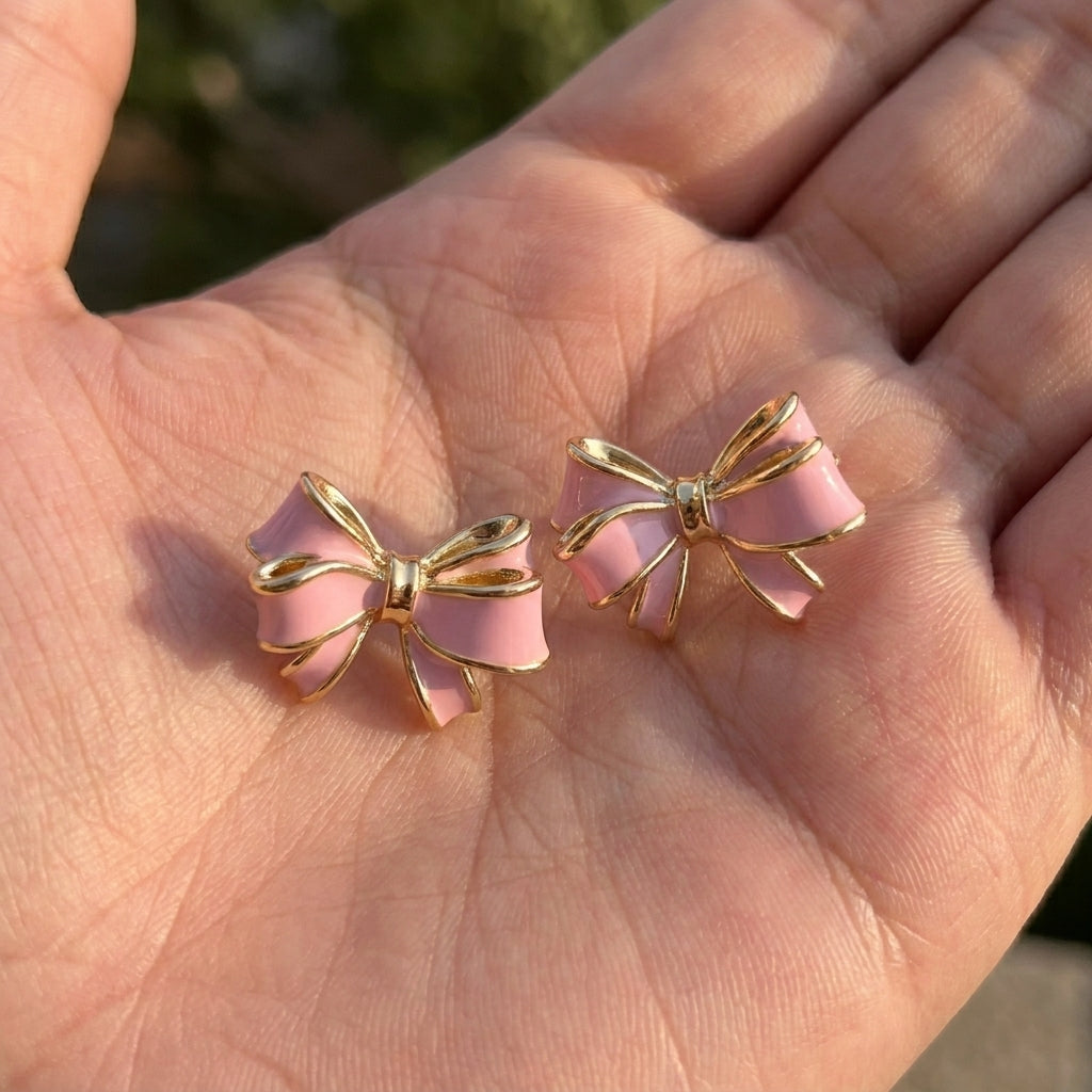 Close-up of a hand holding the pink enamel and gold bow stud earrings under sunny natural daylight.