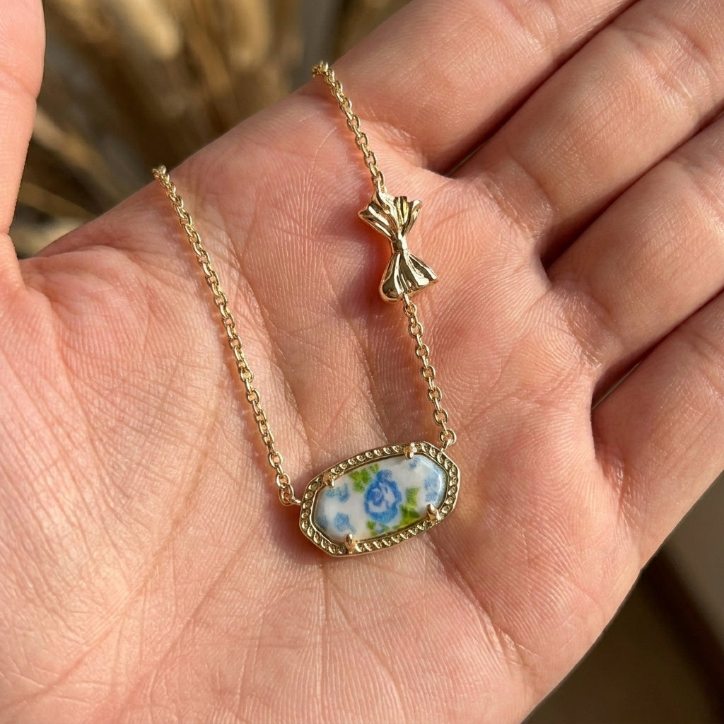 Close-up of the Kendra Scott x LoveShackFancy blue rose floral ribbon bow necklace held in a hand for quality and scale reference.
