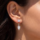 A close-up of a person's ear wearing a silver drop earring with a round stud and a dangling pear-cut clear gemstone.