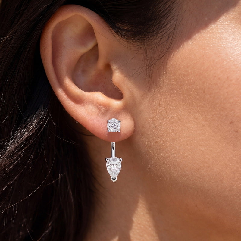 A close-up of a person's ear wearing a silver drop earring with a round stud and a dangling pear-cut clear gemstone.