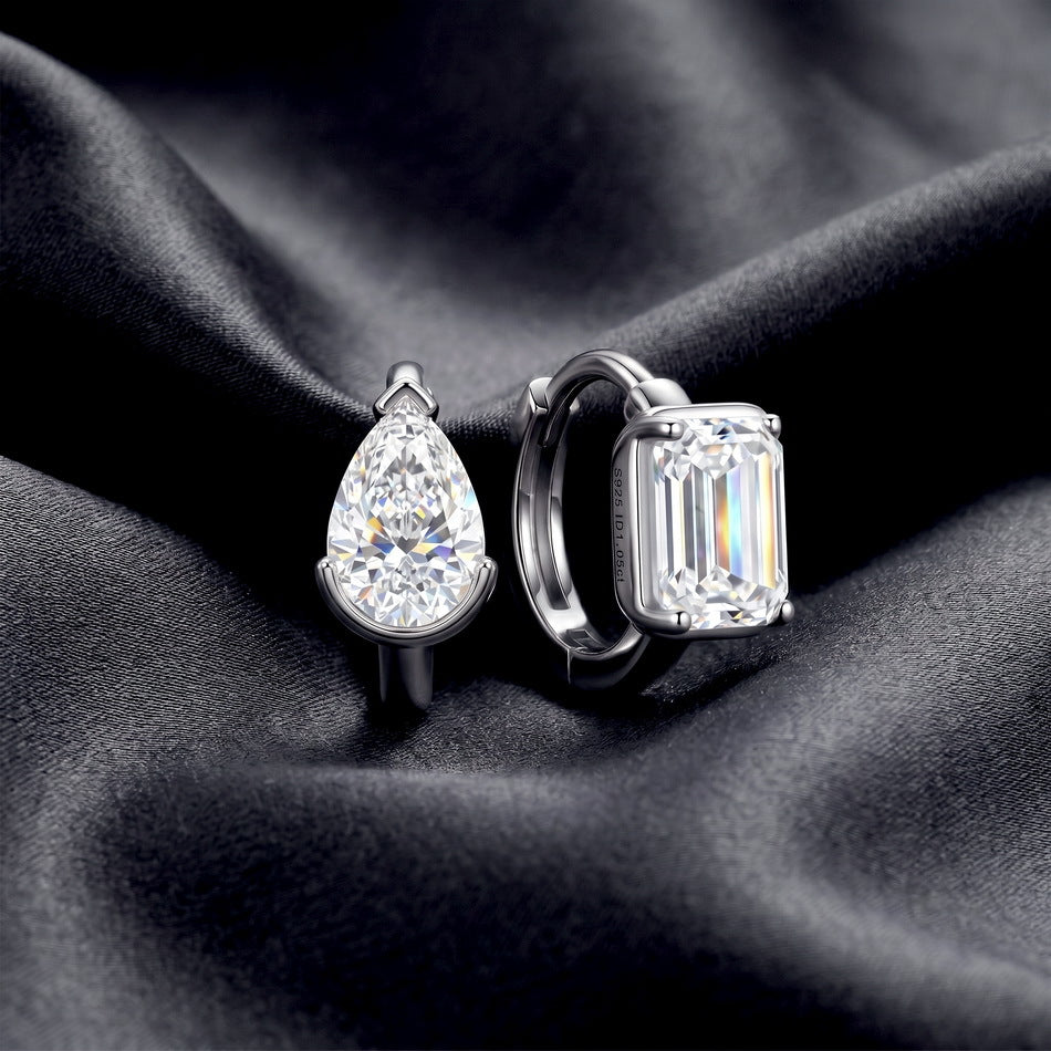 Pair of mismatched pear and emerald cut lab diamond bezel huggie hoops resting on dark silk fabric.