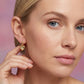 Close-up of a woman wearing a gold heart-shaped earring against a pink background