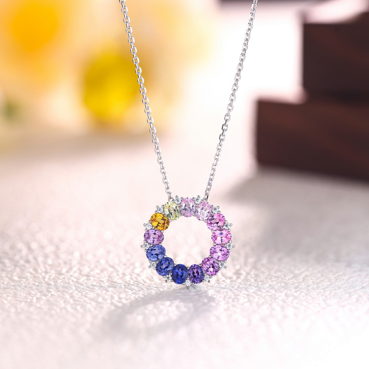 Front angled view of oval cut multi-color gemstone circle pendant resting on white display stage.