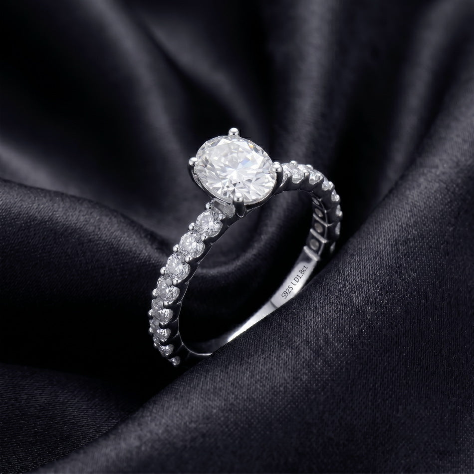 Oval solitaire engagement ring with pave band set against dark silk background.