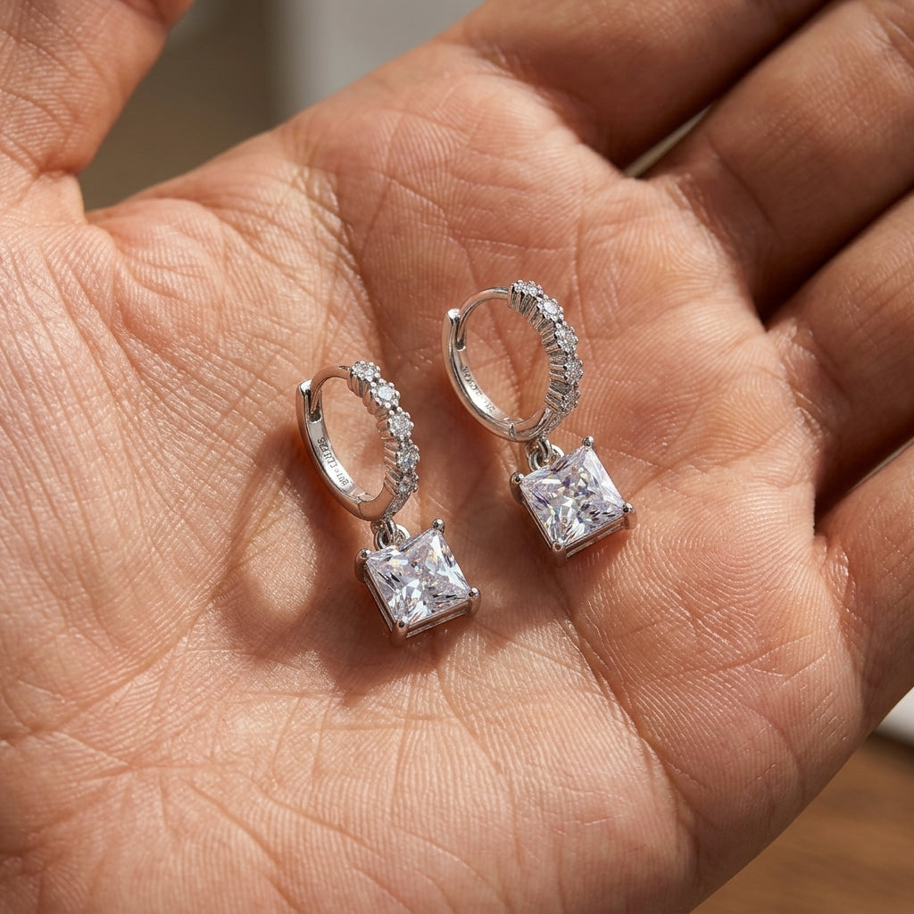 Pair of 2.4ctw princess cut diamond solitaire dangle huggie earrings held in palm.