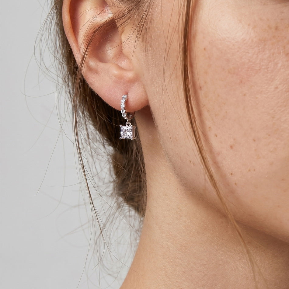 Close-up view of woman wearing princess cut solitaire dangle huggie hoop earring on earlobe.