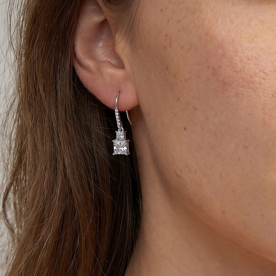 Close-up view of woman wearing princess cut two-stone dangle earrings on earlobe.