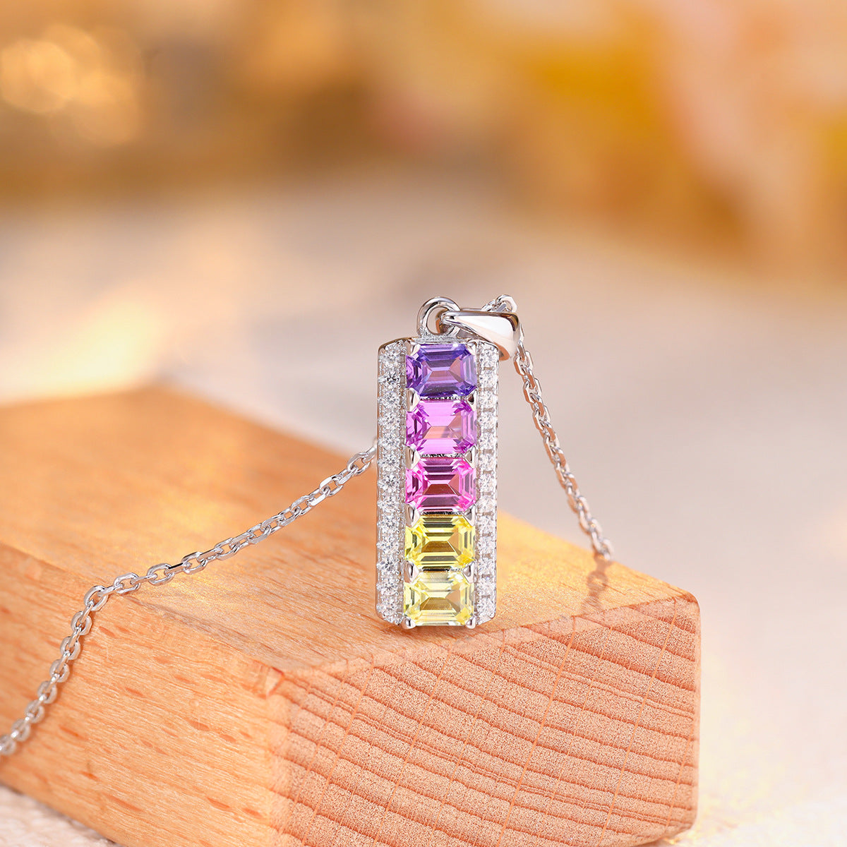 Angled view of rainbow emerald cut gemstone vertical bar pendant resting on wooden display.