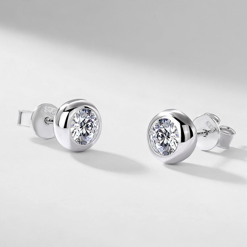 Pair of round bezel set solitaire diamond stud earrings resting on light gray background.