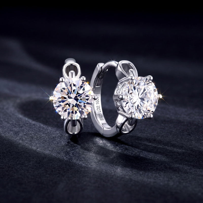 Pair of six-prong round diamond solitaire huggie hoop earrings on black backdrop.
