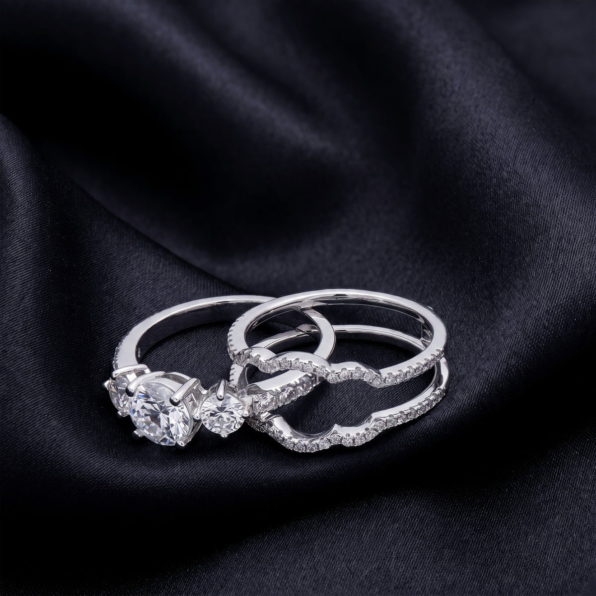 Separated view of 3-stone engagement ring and contour wave enhancer band on dark silk.