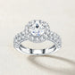 Top studio display of round moissanite halo engagement ring with double row pave accent setting on beige textured background.