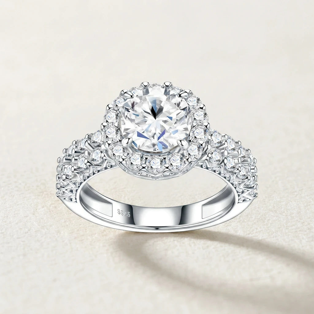 Top studio display of round moissanite halo engagement ring with double row pave accent setting on beige textured background.