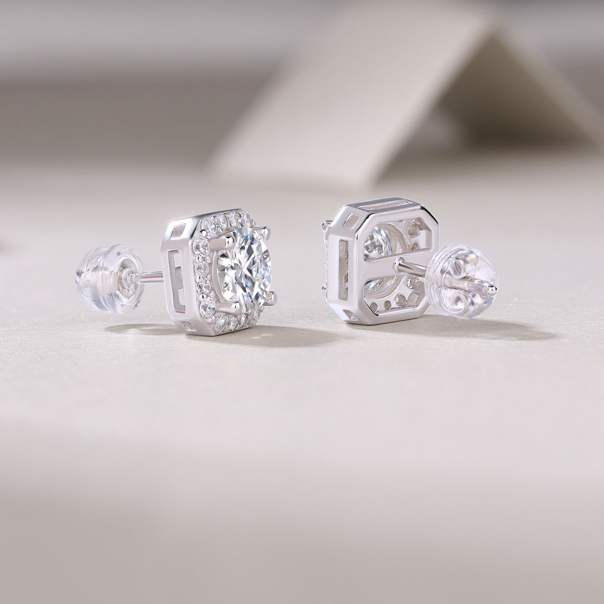 Pair of round cut moissanite octagon cushion halo stud earrings resting on textured surface.