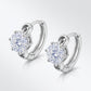 Angled pair of six-prong round diamond solitaire huggie hoops on light grey gradient.