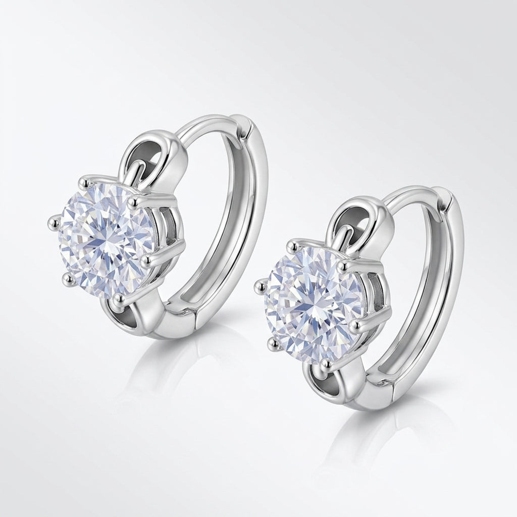 Angled pair of six-prong round diamond solitaire huggie hoops on light grey gradient.
