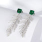 Side angle studio view of square emerald wheat tassel statement earrings resting on a white geometric prop.