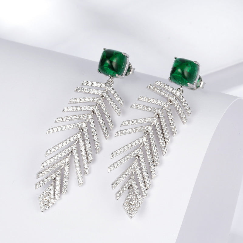 Side angle studio view of square emerald wheat tassel statement earrings resting on a white geometric prop.