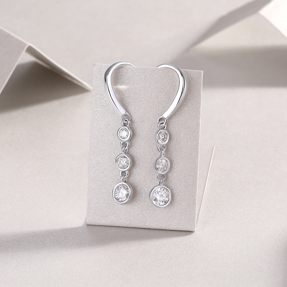 Graduated bezel set moissanite linear drop earrings displayed on grey paper card.