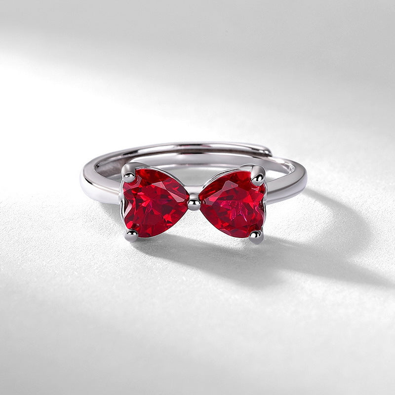Front angle studio view of a vintage heart-shaped ruby bow-knot open ring resting on a shaded white surface.